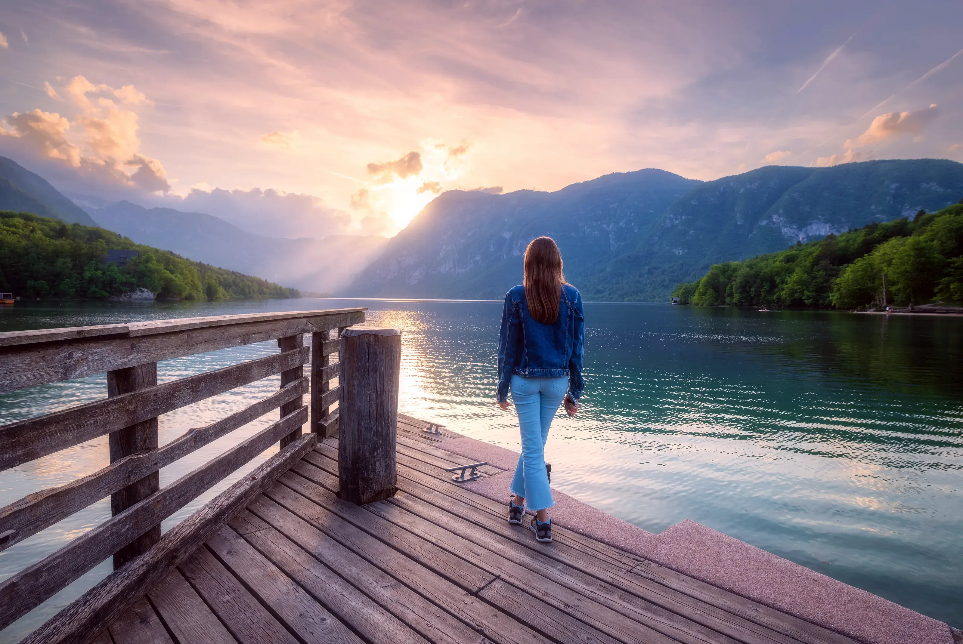 Woman on the wooden pier, mountains and Bohinj lake at sunset in spring. Girl, lake with reflection in water, green forest on mountains and colorful sky in summer.