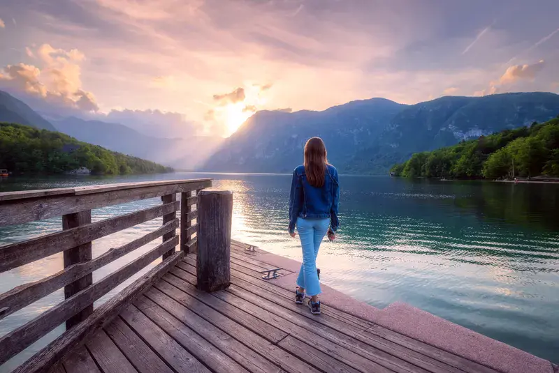 Woman on the wooden pier, mountains and Bohinj lake at sunset in spring. Girl, lake with reflection in water, green forest on mountains and colorful sky in summer.