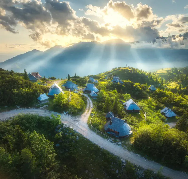 Aerial view of old mountain village and green hills at sunset in summer. Velika Planina, Slovenia. Top view of wooden houses, meadows, rural road, sky, sunbeams, clouds. Alpine shepherds settlement