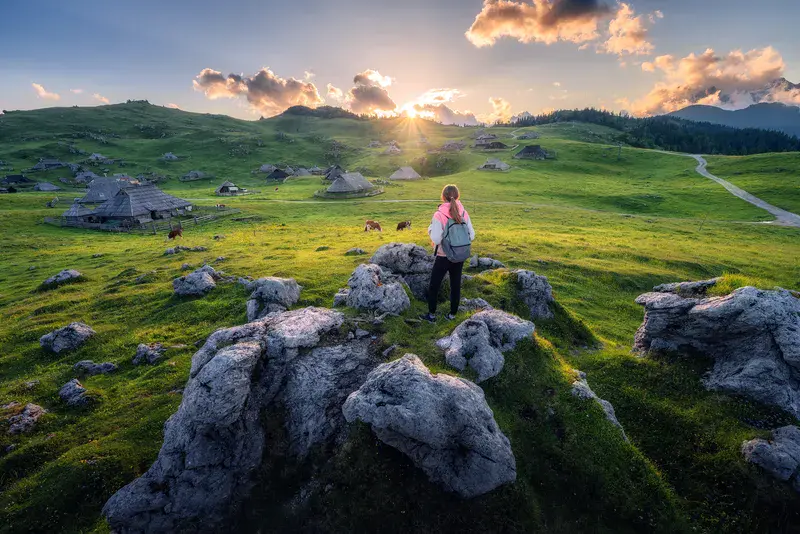 Girl with packpack on the stone, cows on green meadows in beautiful old alpine village in mountains at sunset in summer. Landscape with young woman, houses, trees, sky. Travel and Hiking. Slovenia