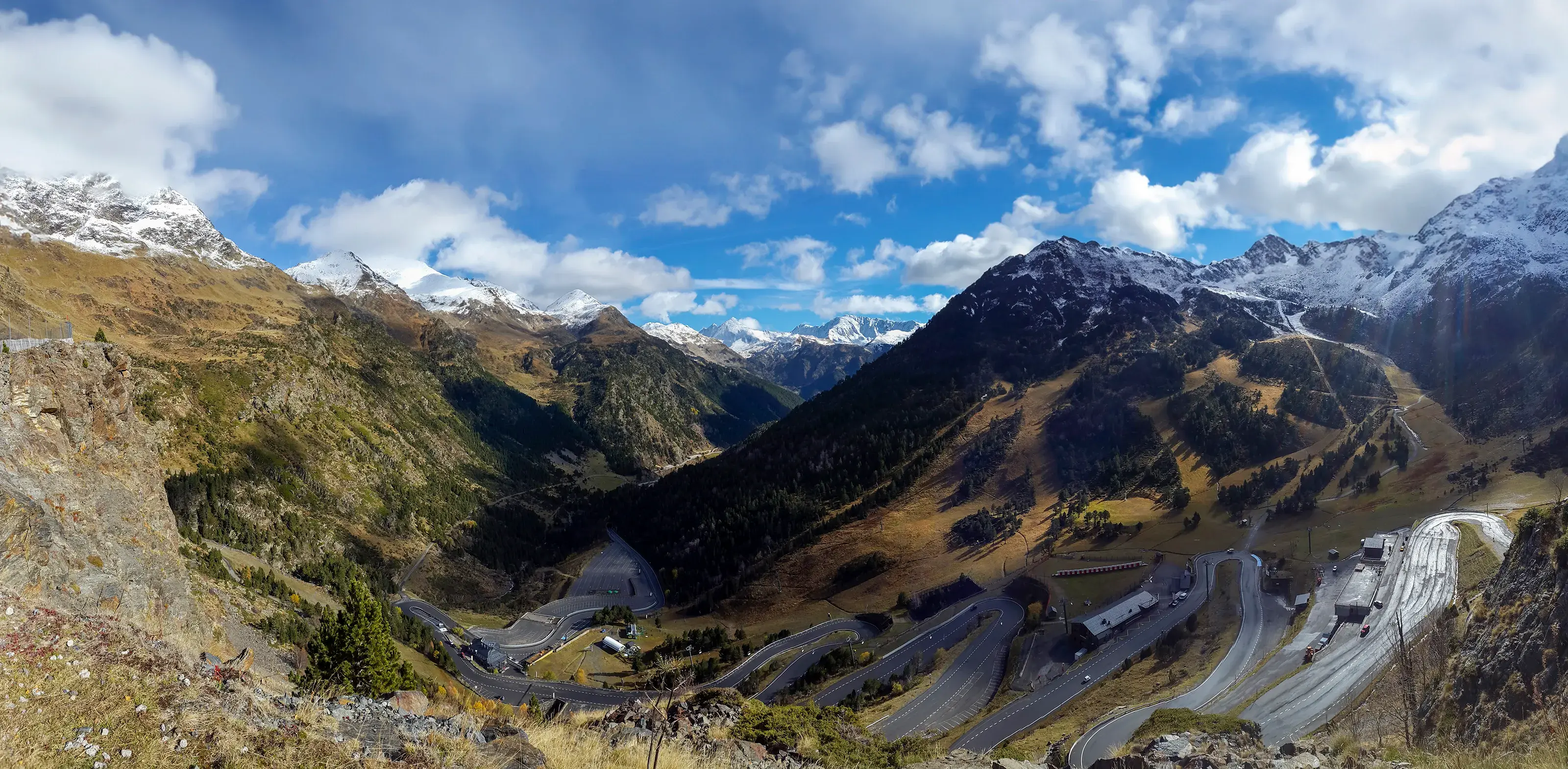 In Andorra, snowy mountain peaks gleam under a blue sky dotted with white clouds. A serpentine road winds its way to a summit, capturing the essence of a sunlit alpine adventure.