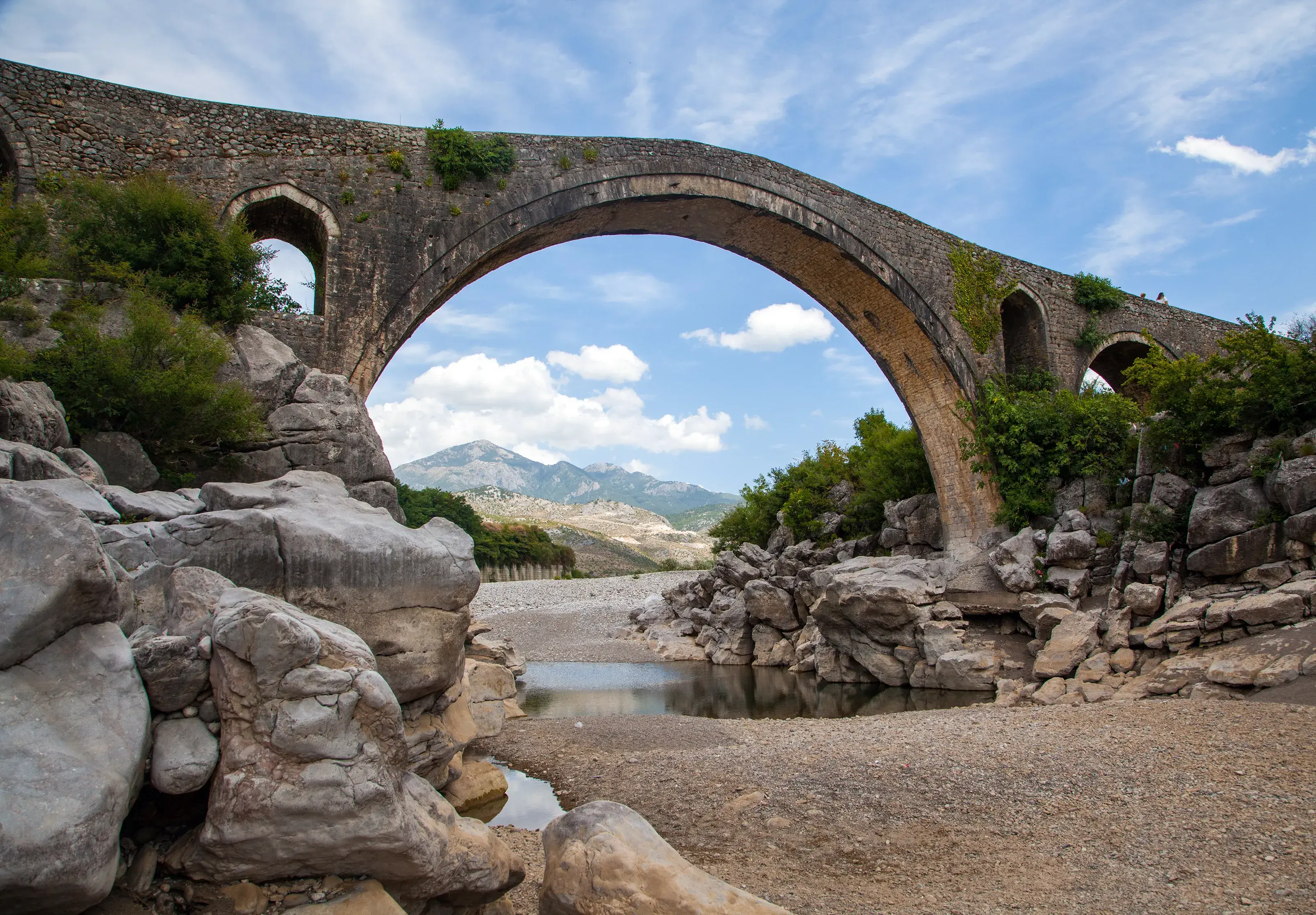 An ancient arched stone bridge in Bulgaria spanning a rocky, dry riverbed with green mountains in the background under a blue sky.