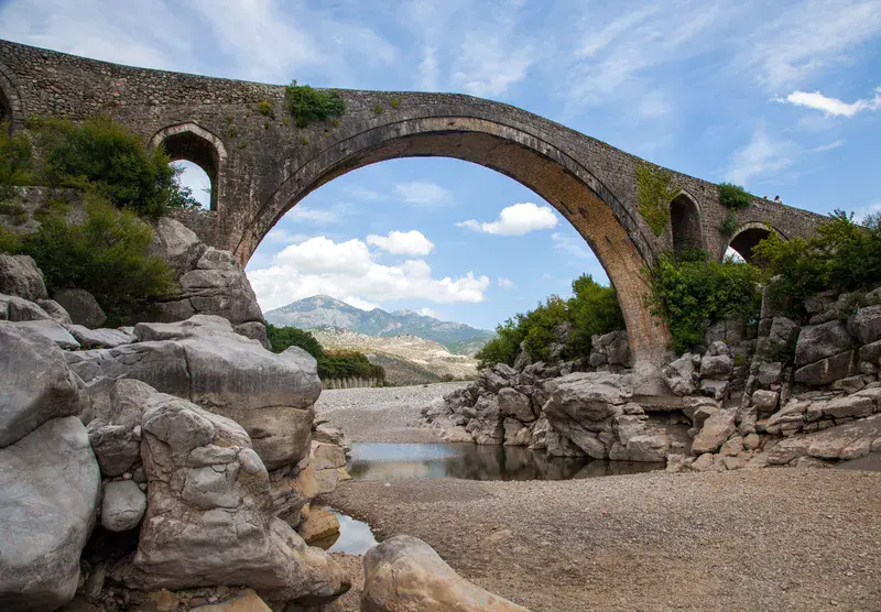 An ancient arched stone bridge in Bulgaria spanning a rocky, dry riverbed with green mountains in the background under a blue sky.