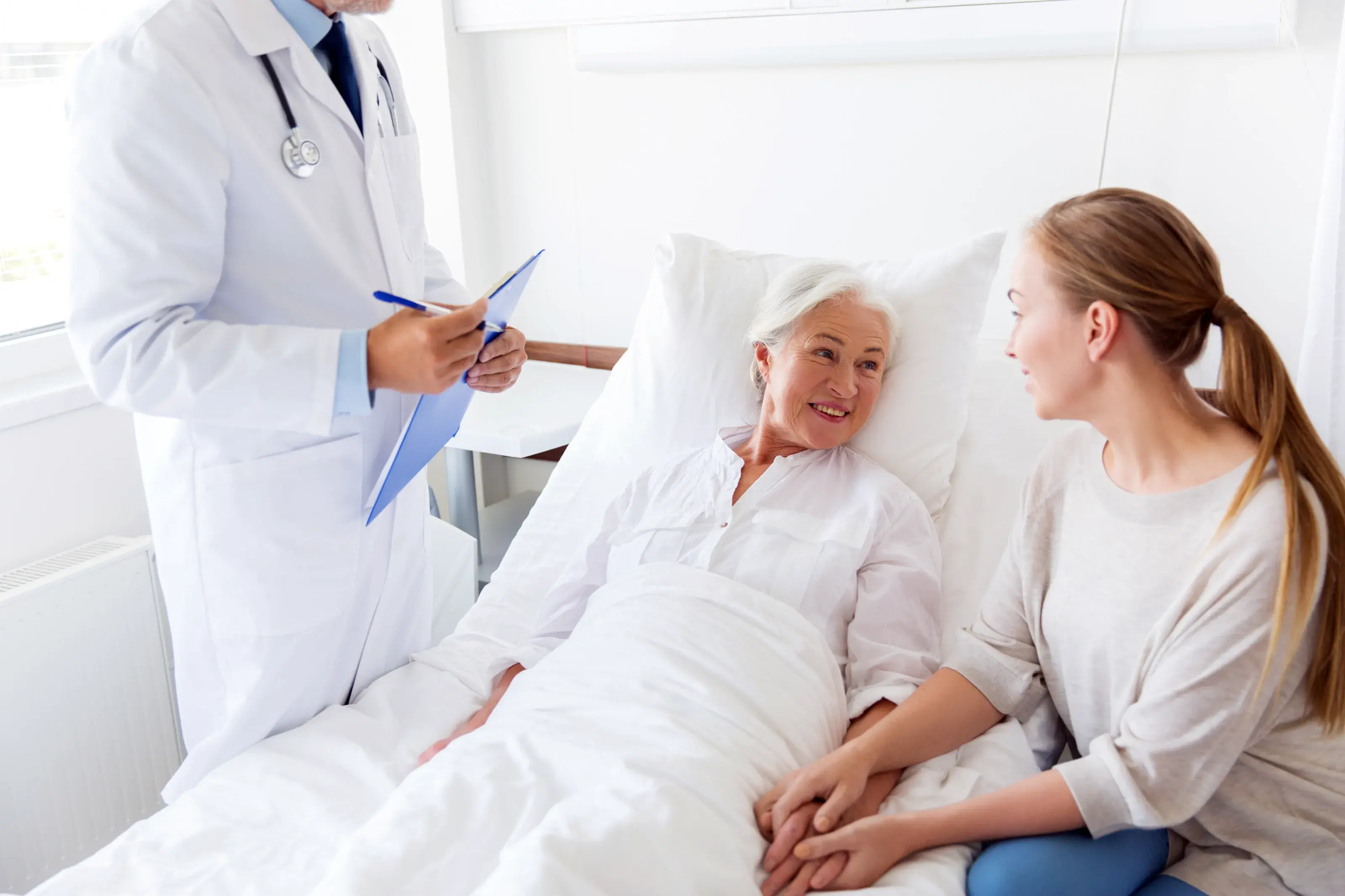 A doctor in a white coat speaks with a smiling senior female patient in a hospital bed, as a younger woman, likely a daughter, holds her hand.