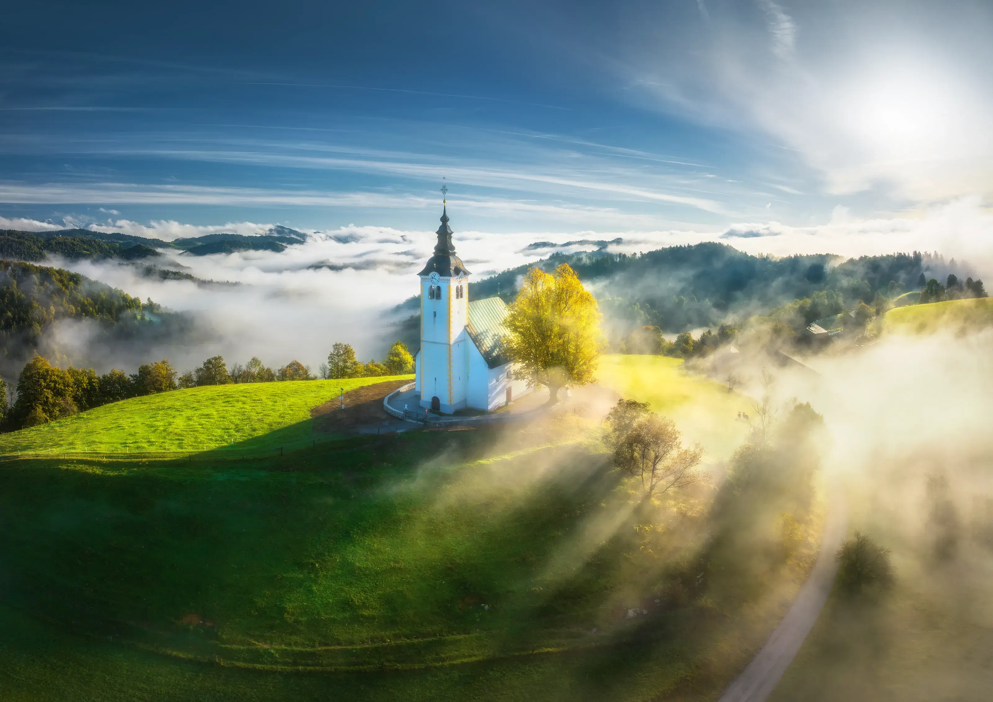 The iconic St. Primus and Felician Church in Jamnik, Slovenia, perched on a vibrant green hill and surrounded by a sea of morning fog with sunbeams breaking through.