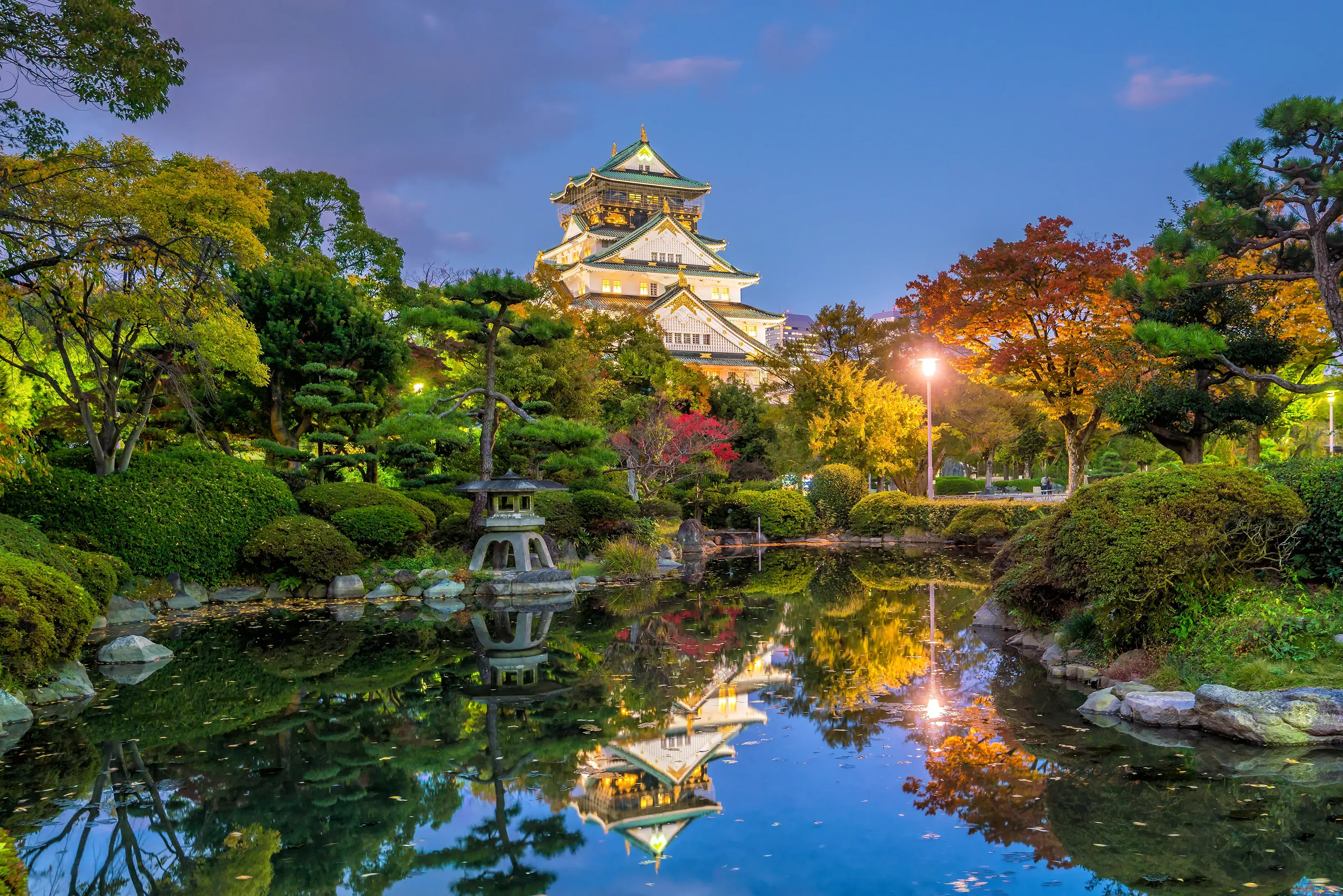 The historic Osaka Castle illuminated at dusk, perfectly reflected in the calm waters of its surrounding moat, with autumn foliage.