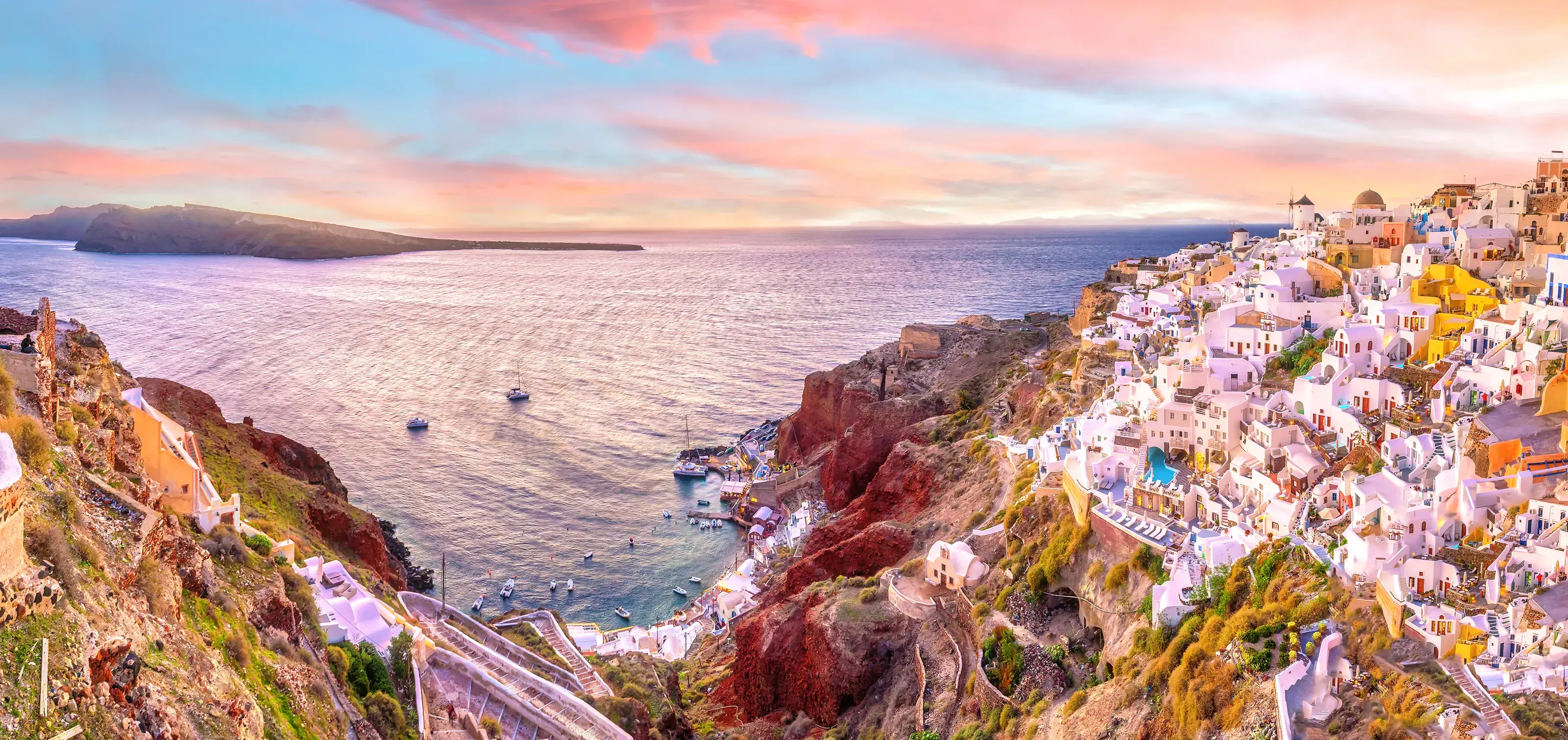 A stunning panoramic sunset view of the village of Oia in Santorini, Greece, with its iconic white buildings and windmills overlooking the Aegean Sea.