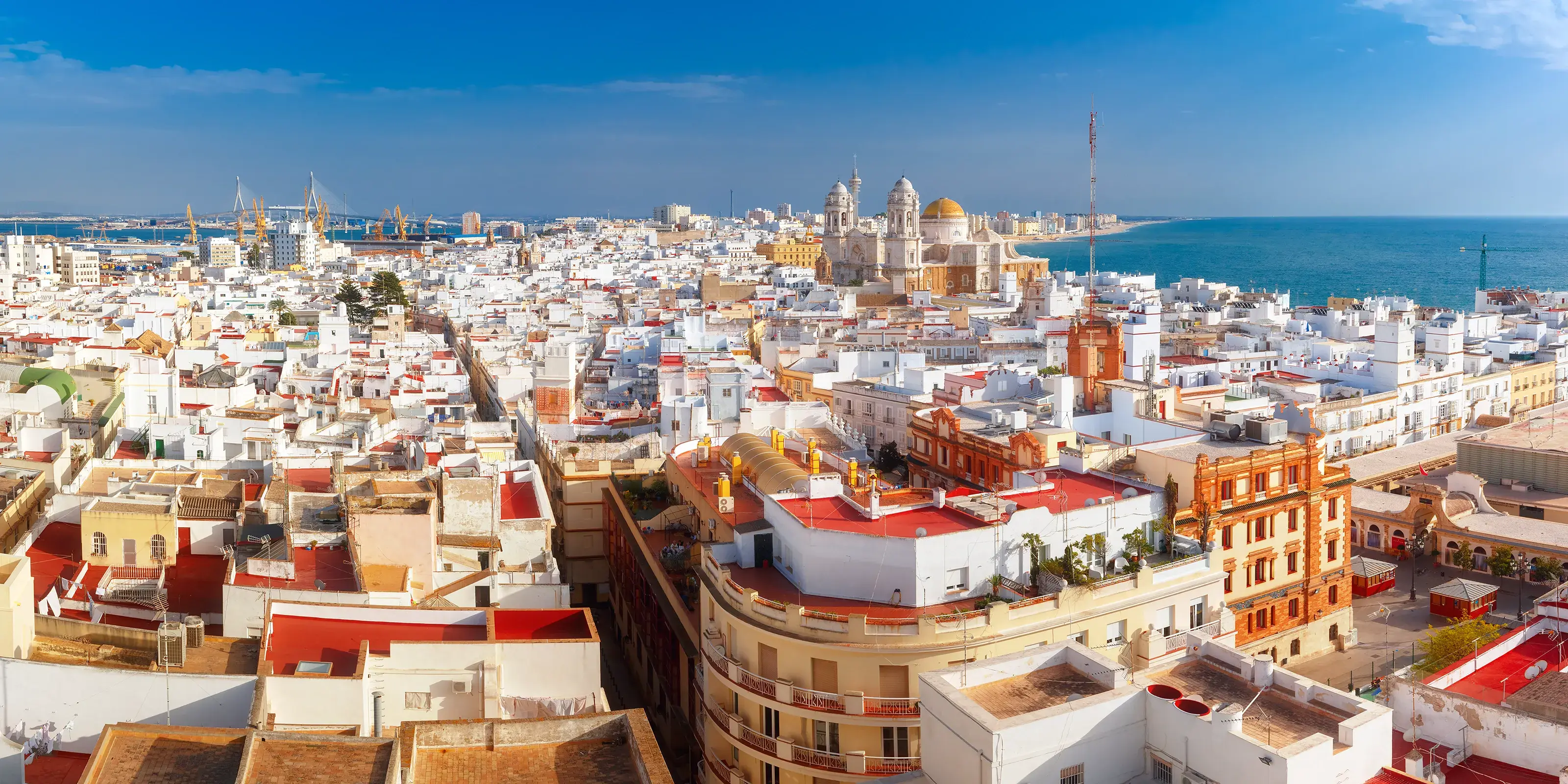 A beautiful panoramic aerial view of the historic city of Cadiz, Spain, showcasing its dense white buildings and the golden dome of the Cadiz Cathedral against the blue sea.