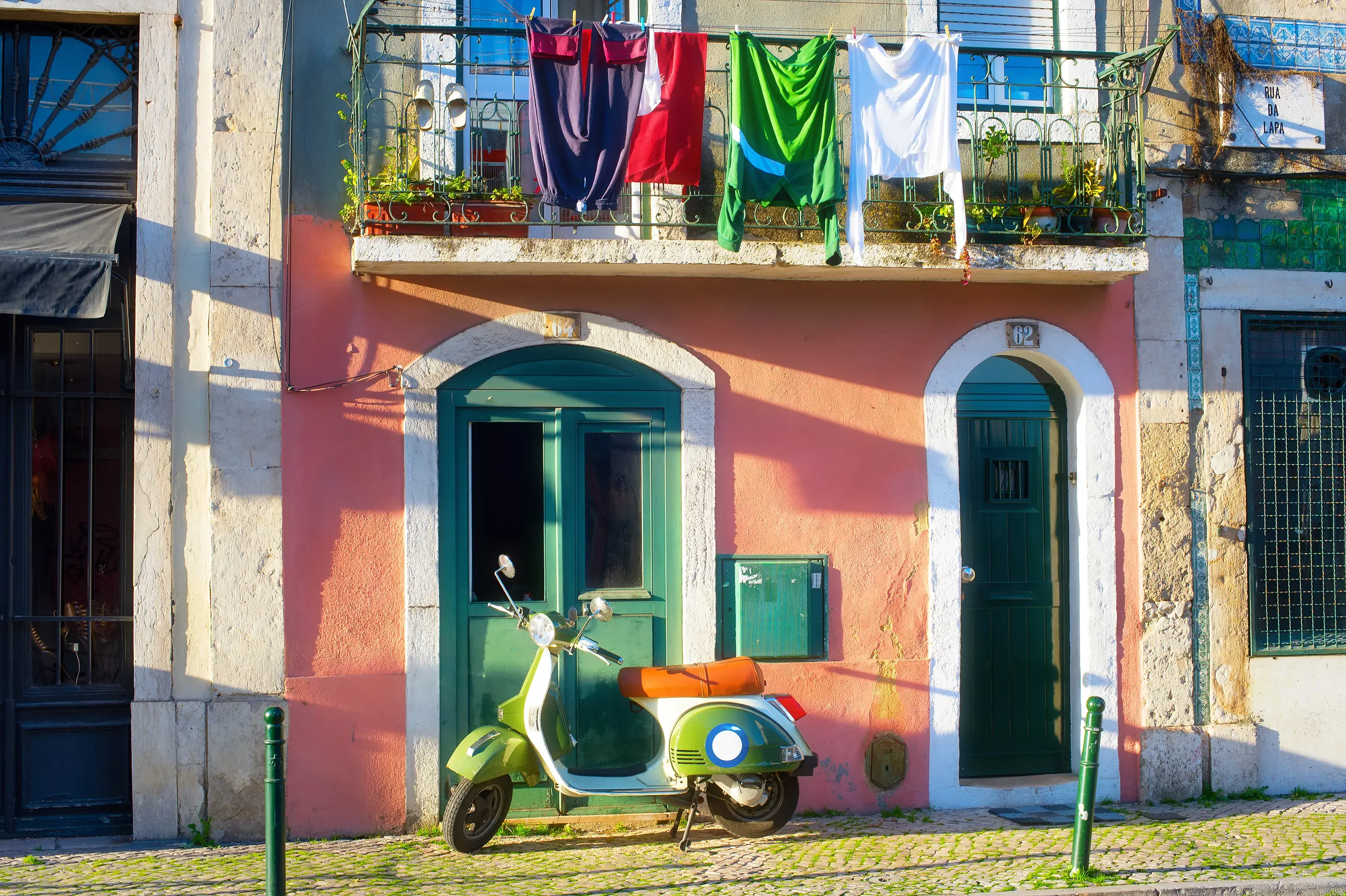 A charming Portugal street scene featuring a classic green scooter parked on a cobblestone sidewalk in front of a rustic pink building with laundry hanging out to dry.