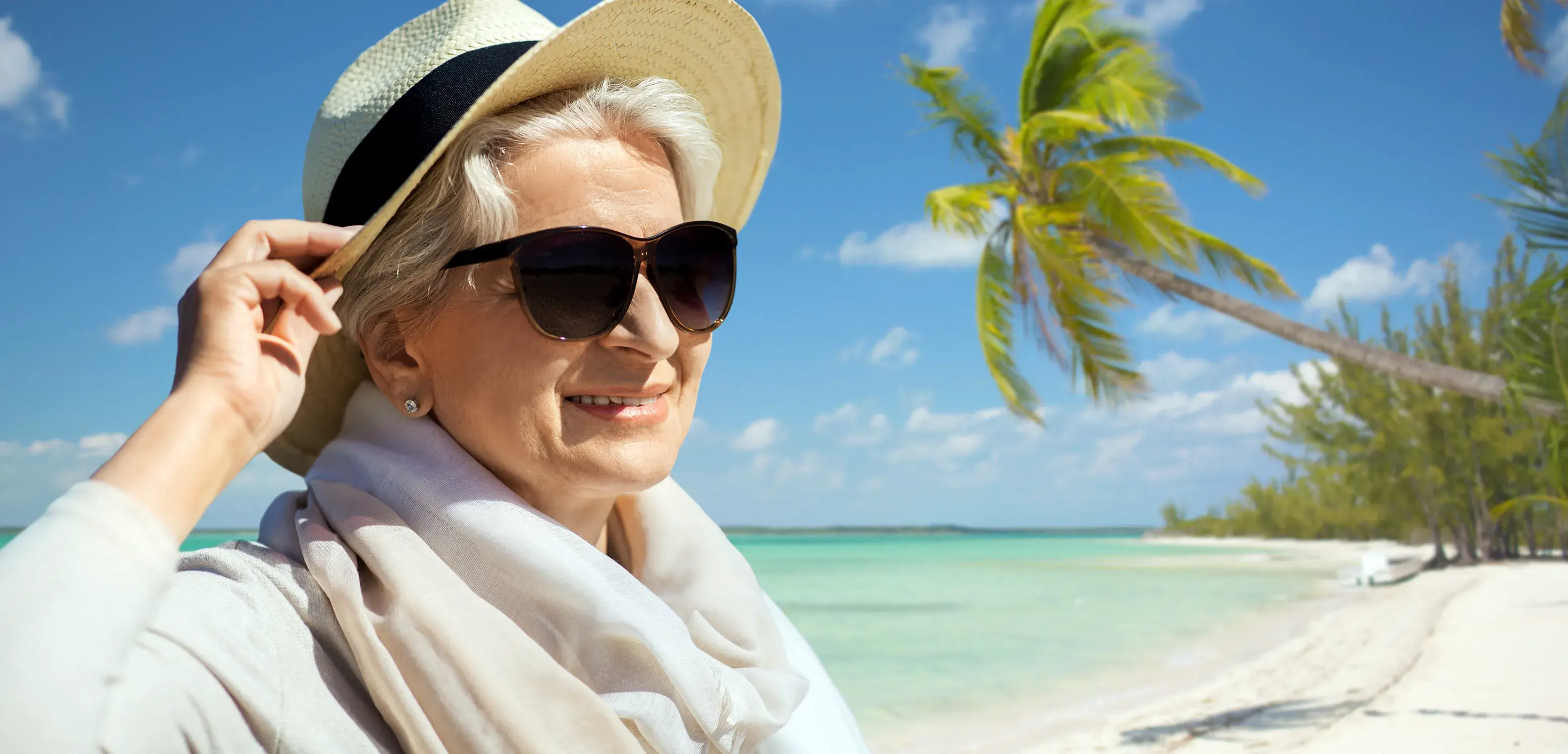 A stylish senior woman wearing sunglasses and a straw hat, smiling contentedly on a beautiful tropical beach with a palm tree and turquoise water.