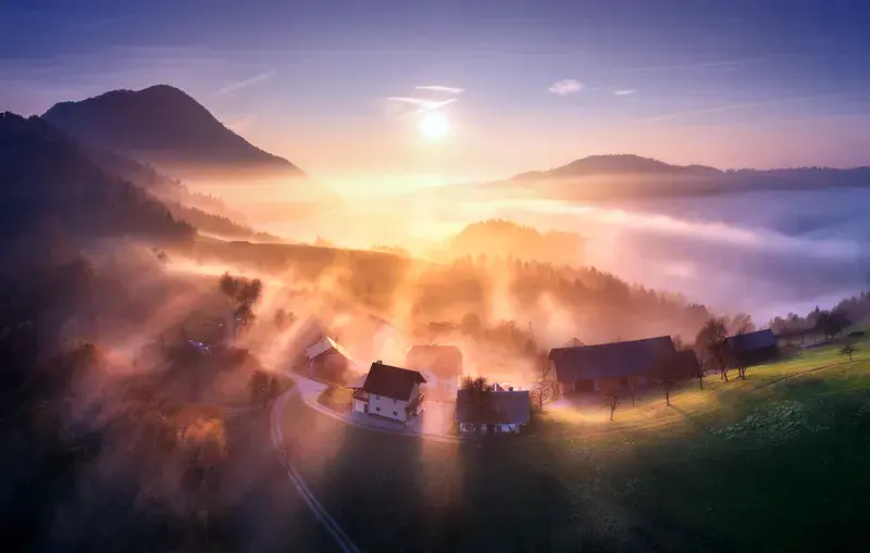 Aerial view of mountain village in orange low clouds at sunrise in autumn in Slovenia. Top view of beautiful houses on hill in fog, green meadows, trees, sky at golden dawn in fall. Colorful landscape