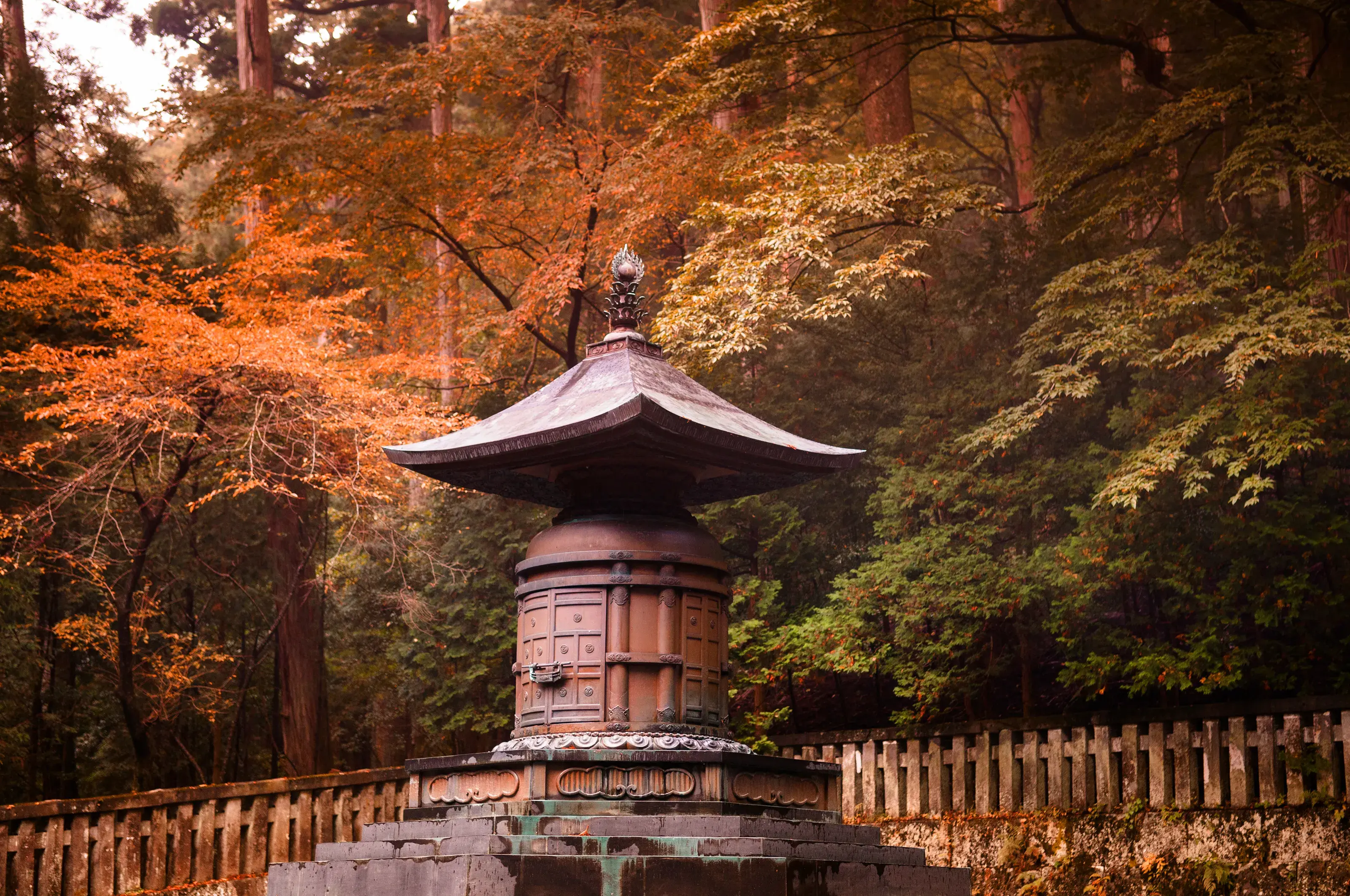 A traditional bronze Japanese pagoda lantern standing in a serene forest clearing, surrounded by vibrant orange and red autumn leaves.