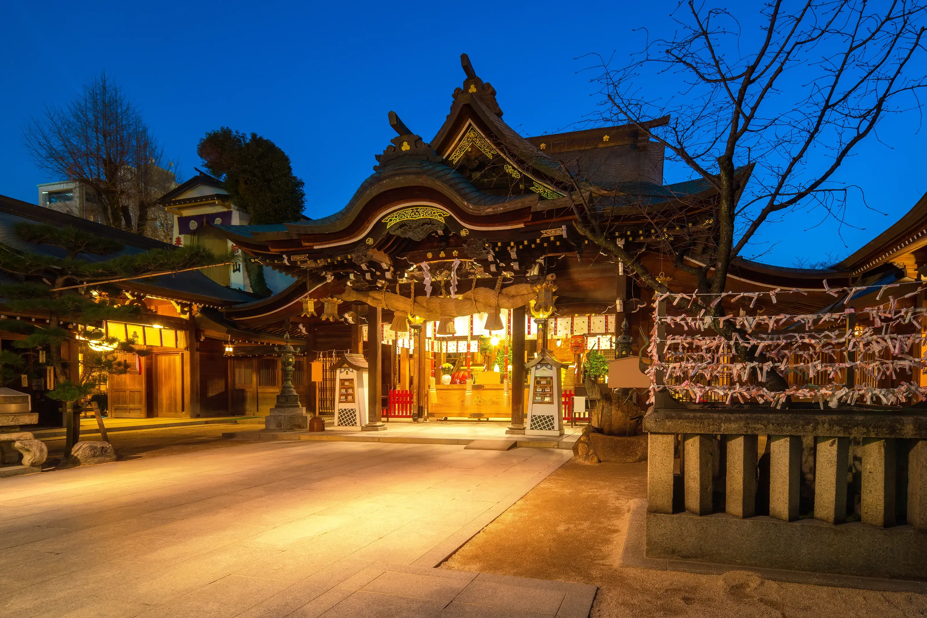 A beautifully illuminated traditional Japanese shrine at night, with intricate wooden architecture and rows of omikuji paper fortunes tied to a frame.